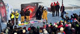 Zdjęcie przedstawia grupę funkcjonariuszy i przedstawicieli służb stojących w jednym szeregu przy zamarzniętym jeziorze. Wśród nich widoczni są strażacy Państwowej Straży Pożarnej w umundurowaniu służbowym, policjanci oraz przedstawiciele innych służb. Strażak trzyma mikrofon i przemawia do zgromadzonych uczestników. W tle znajdują się banery informacyjne Państwowej Straży Pożarnej z hasłem „Zgaś ryzyko zapal świadomość”. Zdjęcie ukazuje oficjalne rozpoczęcie wydarzenia plenerowego.