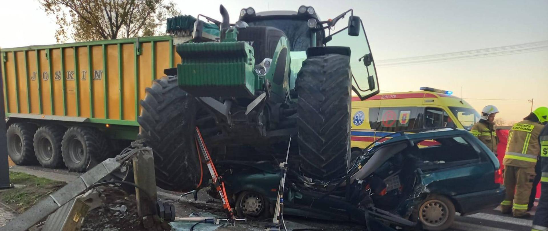 Ciągnik rolniczy najechał kołami na samochód osobowy. Widoczny zmiażdżony samochód. Rozłożone narzędzia hydrauliczne. W tle karetka pogotowia i stojący przy niej strażacy w ubraniach specjalnych. 