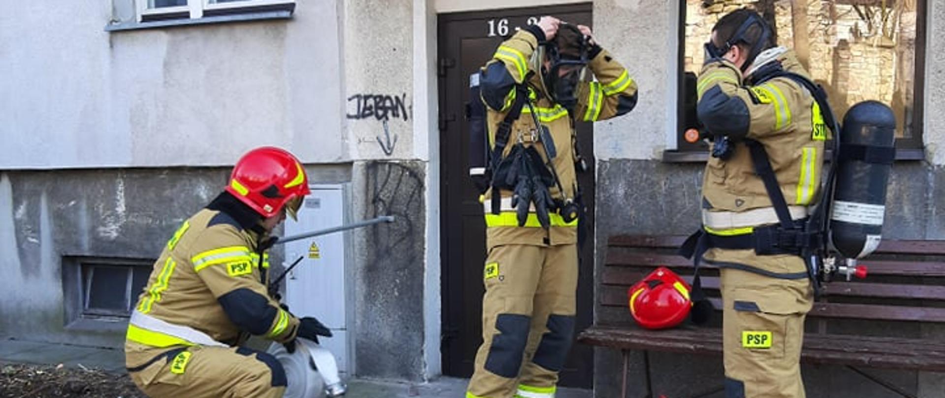 Dwaj strażacy w ubraniach specjalnych przygotowują się do wejścia na klatkę schodową zakładając sprzęt ochrony dróg oddechowych. Obok strażak buduje linię gaśniczą