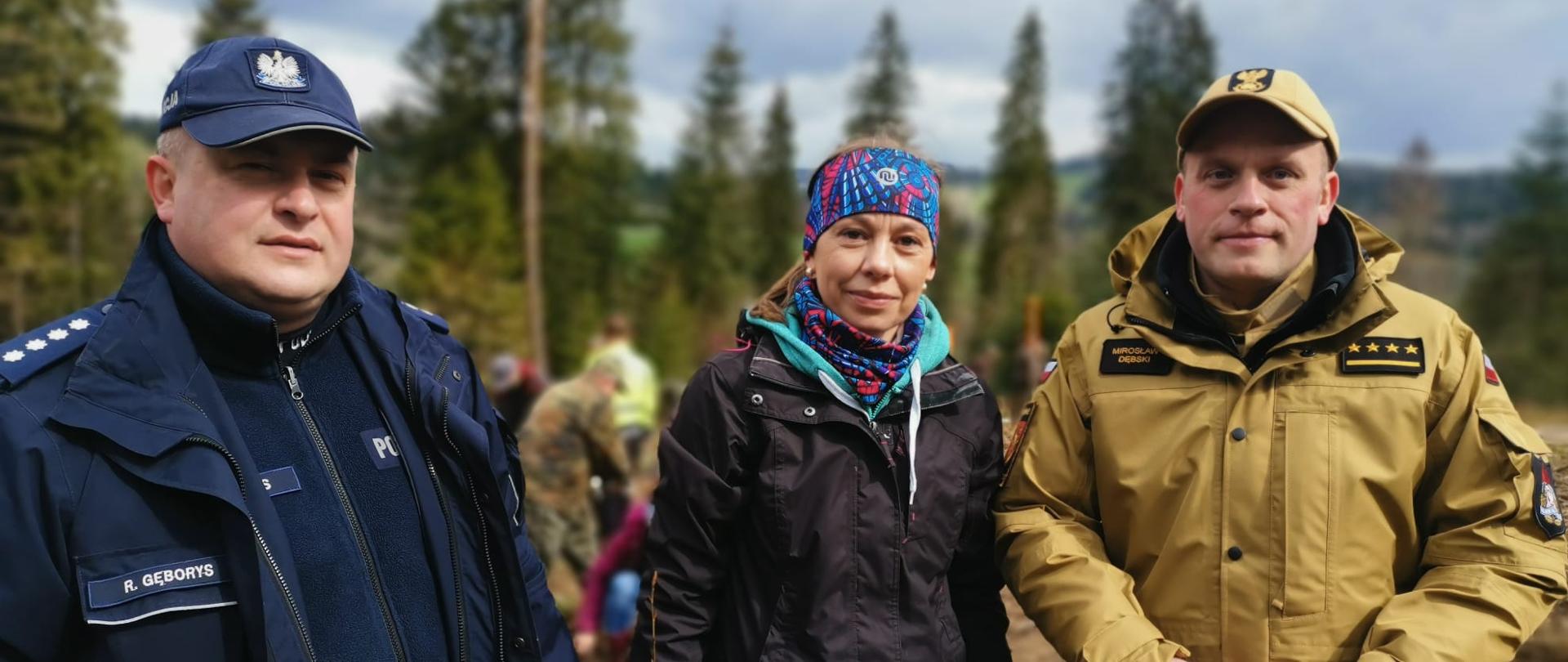 Na zdjęciu widać zastępce dowódcy jednostki ratowniczo gaśniczej PSP w Ustrzykach Dolnych oraz kobietę i Policjanta. W tle las oraz ludzie którzy sadzą sadzonki.