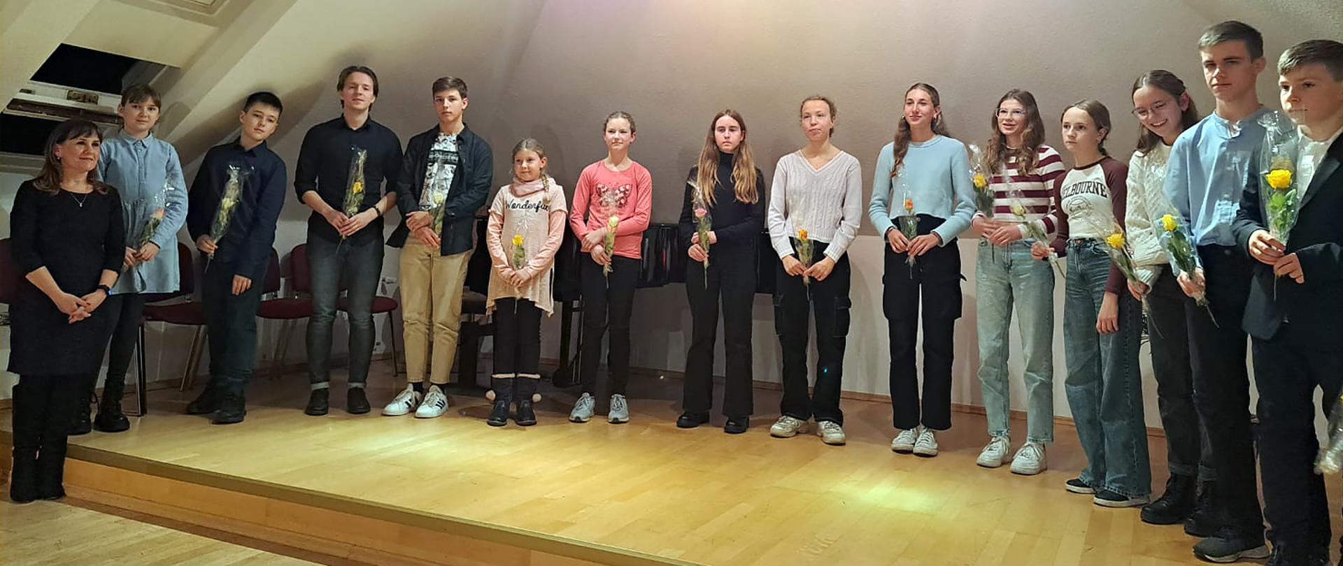 Na zdjęciu na tle jasnej ściany, w szeregu stoi grupa 15 osób, jedna osoba dorosła i 14 dzieci, dzieci trzymają w rękach żółte kwiatki. Wszyscy twarzami zwróceni w kierunku fotografującego.