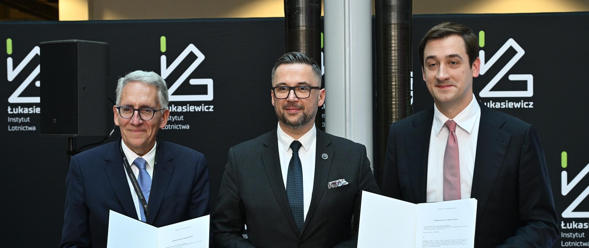 Na tle czarnej ścianki z napisem Sieć Badawcza Łukasiewicz stoi trzech mężczyzn w garniturach, pośrodku minister Kulasek, pozostali pokazują trzymane w rękach otwarte teczki z dokumentami.
