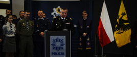 Strażak stojąc przy mównicy przemawia w obecności przedstawicieli służb mundurowych oraz zaproszonych gości za stojącymi osobami na ścianie znajduje się logo Pomorskiej Policji oraz stoją flagi Polski oraz województwa pomorskiego. 