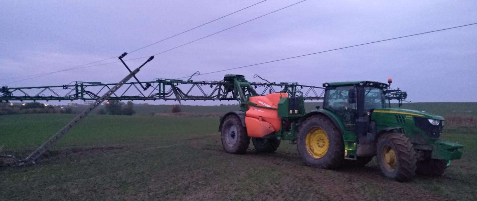 Zdjęcie przedstawia pole, na którym znajduje się ciągnik z opryskiwaczem. Po lewej stronie znajduje się złamany słup energetyczny.