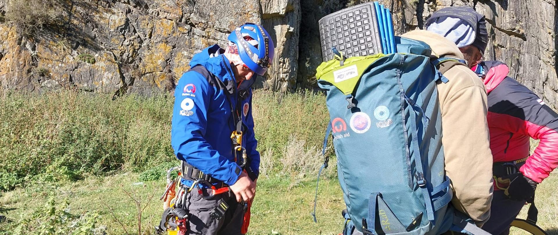 Trzy osoby przygotowują się do wspinaczki, mają na sobie uprzęże do wspinaczki, plecaki, a tle widać stromą ścianę