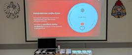 Stół z zestawami czujek dymu i materiałami demonstracyjnymi ustawiony przed ekranem projekcyjnym. Na ekranie slajd informacyjny wyjaśniający działanie autonomicznej czujki dymu, w sali szkoleniowej PSP.