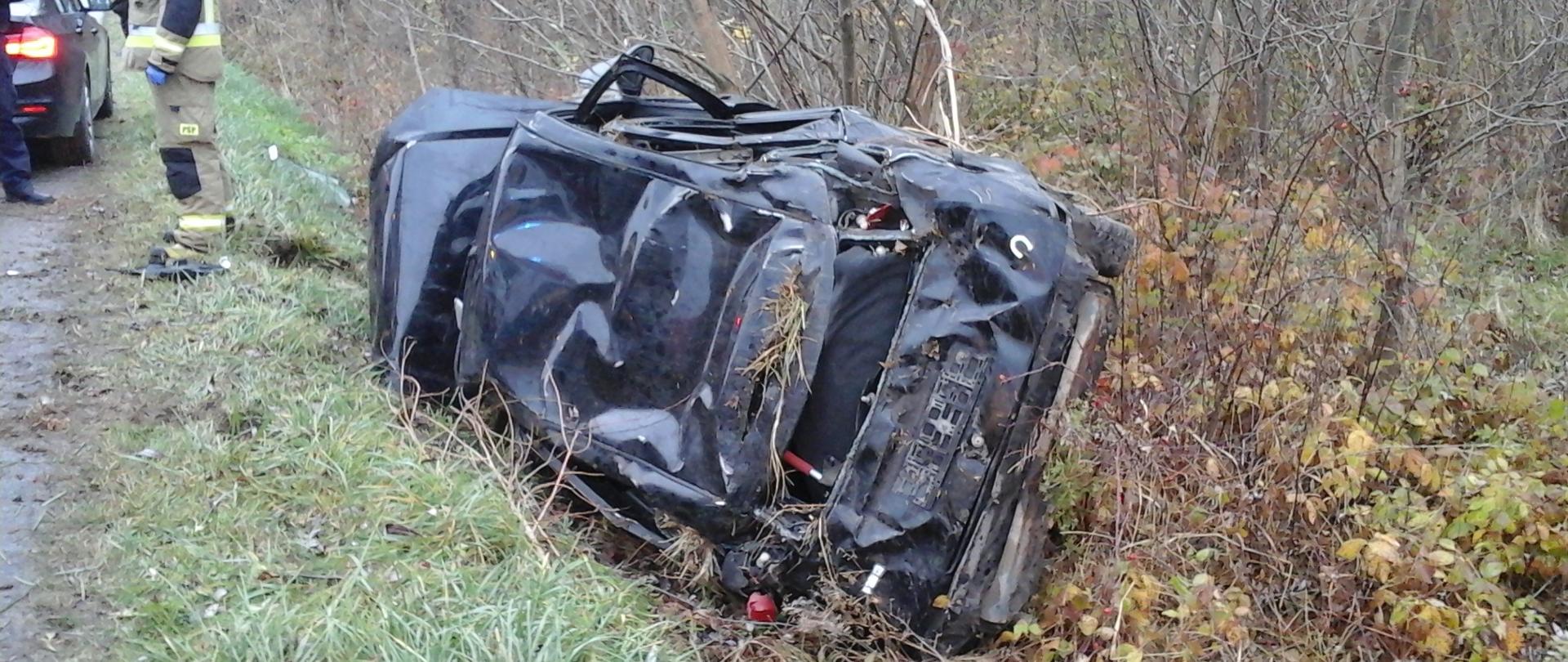 Uszkodzony samochód osobowy leżący na boku w przydrożnym rowie.