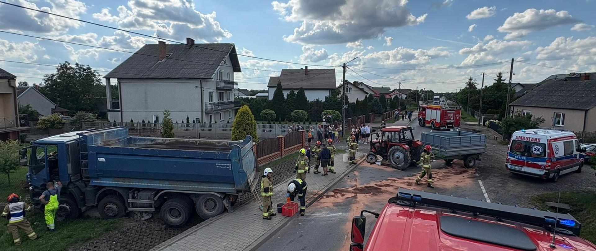 Rozbity ciągnik rolniczy z przyczepą na środku jezdni, plama oleju zabezpieczana przez strażaków w umundurowaniu bojowym, z prawej strony karetka pogotowia, z lewej ciężarówka, w oddali widać budynki mieszkalne i pojazdy strażackie. 