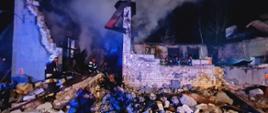 Zdjęcie przestawia funkcjonariuszy Państwowej Straży Pożarnej oraz Ochotniczej Straży Pożarnej podczas działań gaśniczych. W tle pogorzelisko remizy - zniszczony obiekt. 