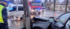 Na zdjęciu widać uszkodzony pojazd osobowy. Uszkodzony bok od kierowcy. Pojazd w kolorze białym. Z tyłu budynki mieszkalne położone przy ulicy Stawiszyńskiej. W środku pojazdy gaśnicze z włączoną sygnalizacją ostrzegawczą. Pojazdy kolor czerwony. Niebo zachmurzone. Pora zimowa. Po lewej widać postać policjanta w mundurze. Między Policjantem, a drugim rozbitym pojazdem w kolorze czarnym drewniany słup telekomunikacyjny.