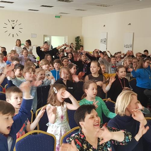 Zdjęcie przedstawia aulę PSM I st. w Grajewie i na niej dzieci tańczące, które uczestniczą w koncercie ,,Dźwięki Wiosny", dzieci są ubrane na kolorowo, mają uniesione dłonie. W tle zegar oraz grafiki z pięciolinią i nutami w kolorach biało- czarnych. 