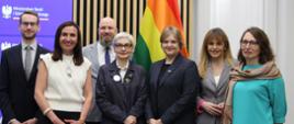 Na tle ściany z cienkich pionowych listewek stoi grupa siedmiu osób, dwóch mężczyzn w garniturach i pięciu kobiet, pośrodku wiceminister Zioło-Pużuk, za nimi z prawej tęczowa flaga.