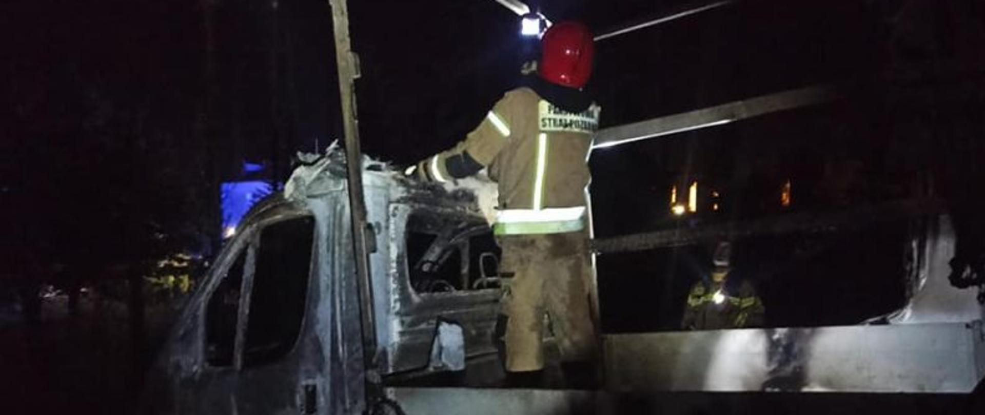 Zdjęcie wykonane w nocy. Tło ciemne, czarne. Oświetlony jest na środku spalony bus i strażak stojący na jego pace. Strażak ma w ręku kamerę termowizyjną. Sprawdza czy pożar został dokładnie ugaszony.
