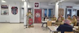 Strażak w mundurze służbowym znajdujące się wewnątrz budynku w dużej sali stoi na mównicy przed mikrofonem. W tle sztandary, godło Polski oraz kilku innych strażaków oraz policjant którzy siedzą przy stolach.