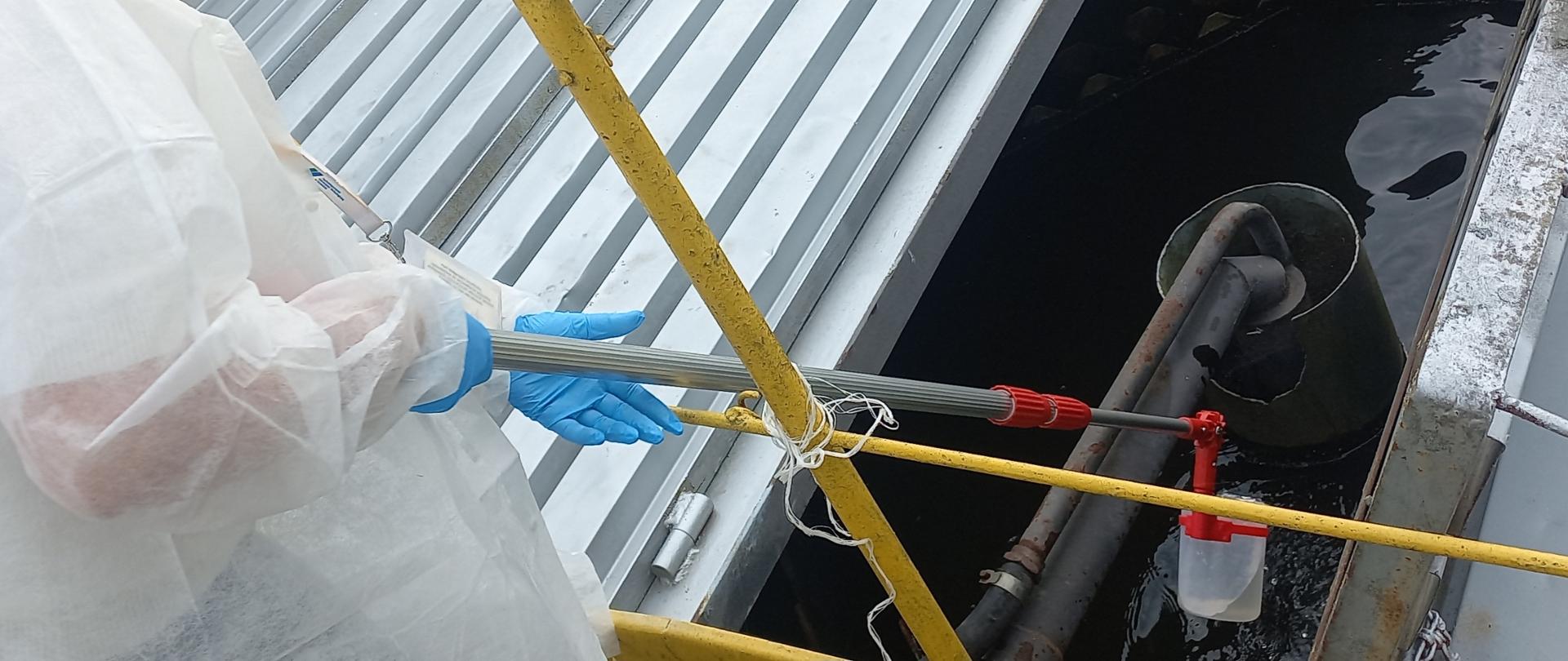 Zdjęcie przedstawia pracownika WIORiN ubranego w odzież ochronną pobierającego próbkę wody ze zbiornika na wodę. 