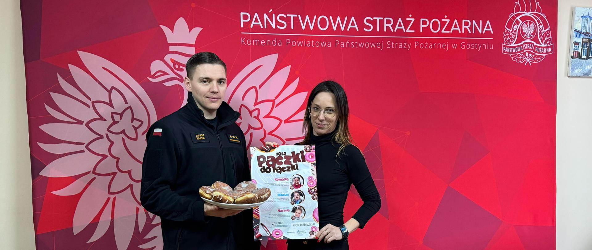 Strażaka oraz kobietę stojących na tle ścianki i pozujących do zdjęcia. Strażak trzyma w ręce talerz pączków. Kobieta trzyma w rękach plakat. na stole przed nimi leżą dwa talerze pączków.