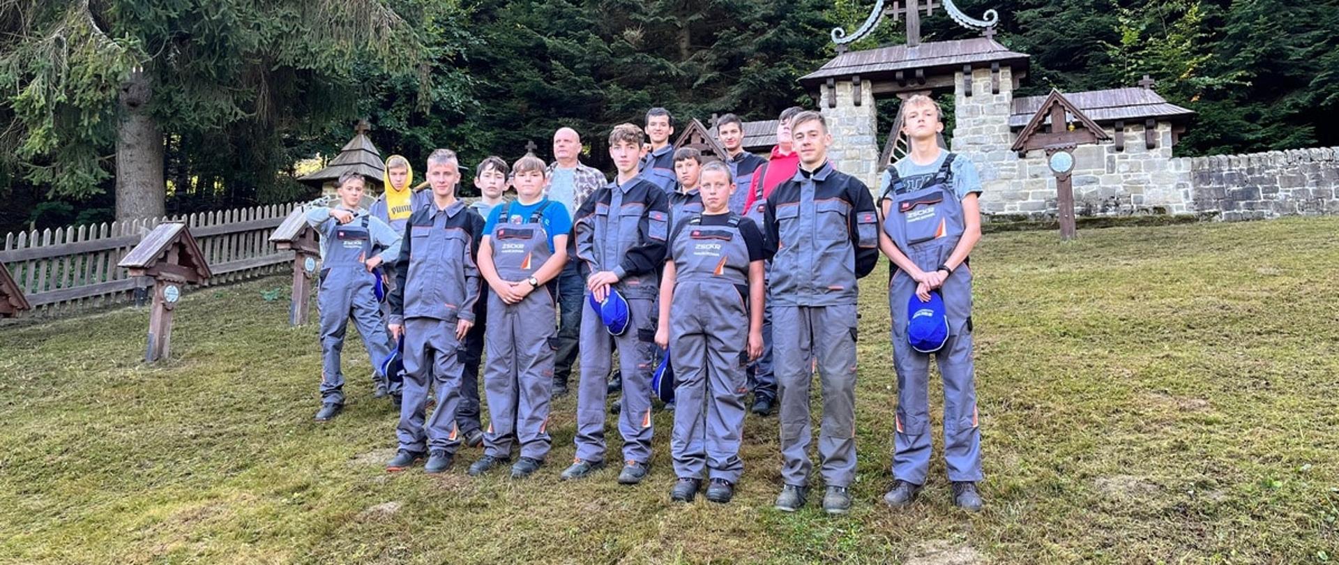 Grupa kilkunastu młodych mężczyzn w szaro-pomarańczowych strojach roboczych pozuje do wspólnego zdjęcia na tle kamiennej bramy i drewnianych krzyży cmentarza wojennego. Teren jest uporządkowany, z krótko skoszoną trawą.