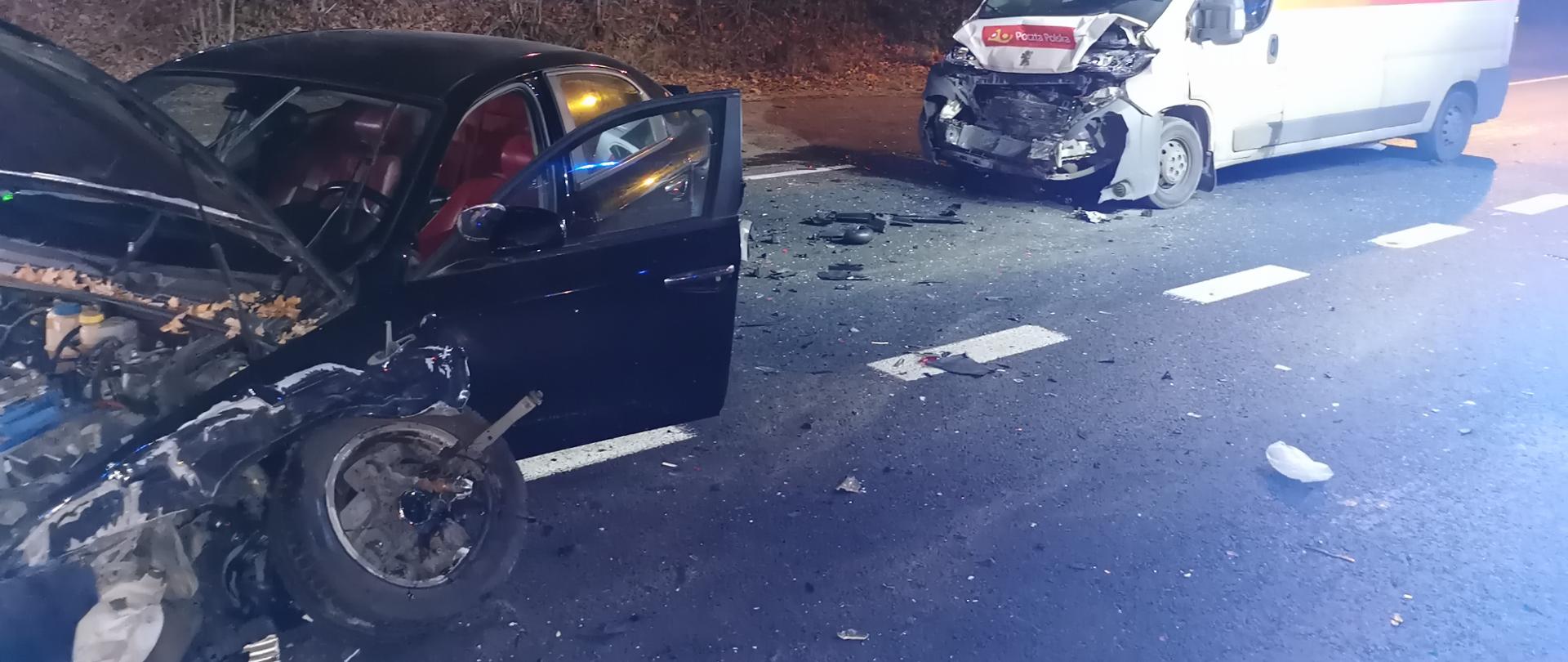 Na zdjęciu widnieją dwa rozbite pojazdy. Po prawej stronie znajduje się biały bus z uszkodzonym przodem. Po lewej stronie znajduje się ciemny samochód osobowy z uszkodzonym przodem, urwanym prawym kołem oraz otwartymi drzwiami i otwartą maską. Pomiędzy pojazdami znajdują się rozsypane części. W tle na poboczu jezdni rosną drzewa.