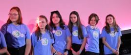 Na tle oświetlonej na różowo-fioletowo ściany stoi sześć młodych dziewczyn w niebieskich koszulkach.