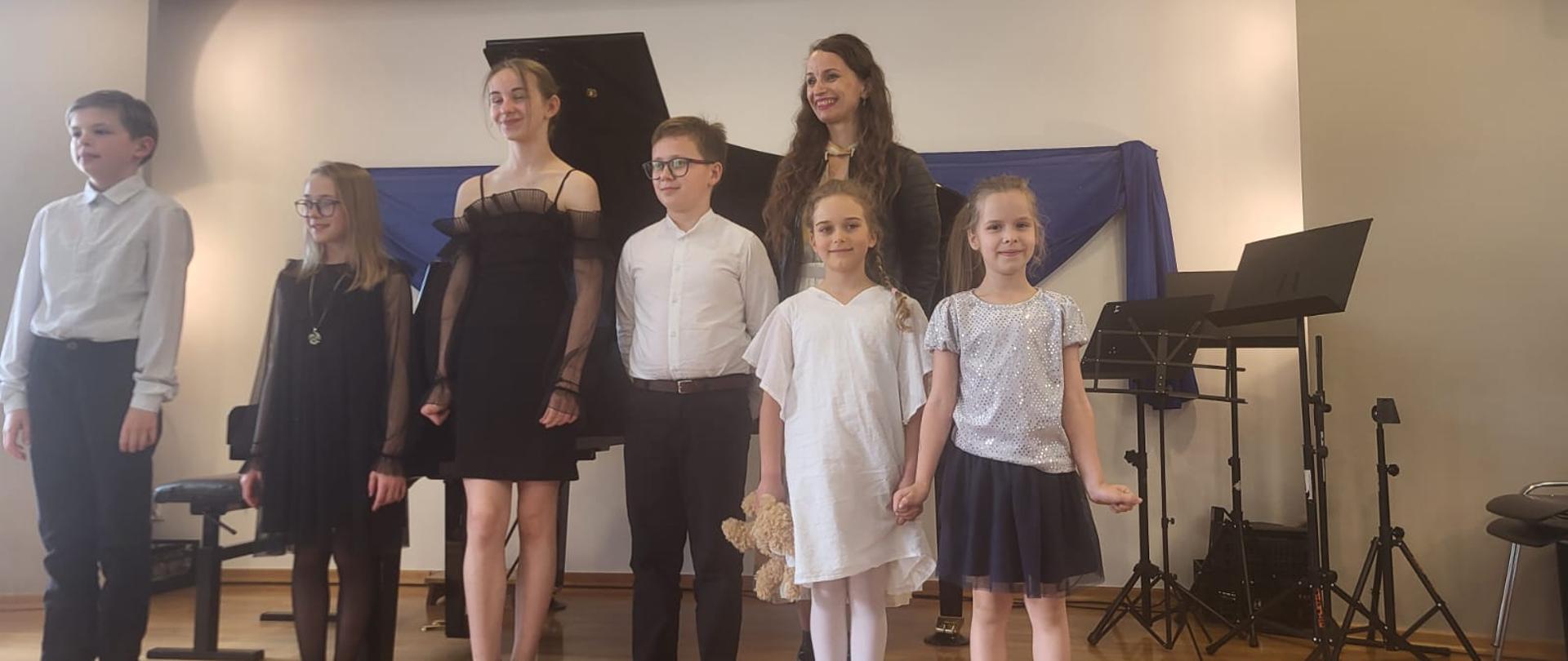 Na zdjęciu widoczna jest grupa dzieci oraz nauczycielka stojący na scenie po zakończonym występie muzycznym. W tle znajduje się fortepian koncertowy. Uczniowie ubrani są elegancko — dziewczynki mają sukienki w jasnych i ciemnych kolorach, a chłopcy białe koszule i ciemne spodnie. Wszyscy stoją obok siebie i patrzą w stronę publiczności. Na pierwszym planie widać fragment widowni. Atmosfera zdjęcia jest uroczysta i radosna, charakterystyczna dla szkolnego koncertu fortepianowego.
