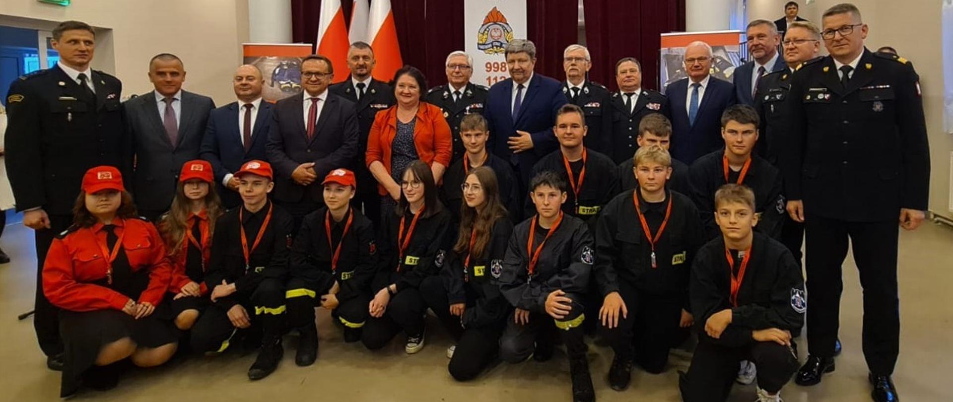 Na zdjęciu widać dzieci z Młodzieżowej Drużyny Pożarniczej pozujących do zdjęcia razem z zastępcą komendanta głównego nadbryg. Krzysztofem Hejdukiem, łódzkim komendantem wojewódzkim PSP oraz zebranymi gośćmi na tle godło i flaga Polski 