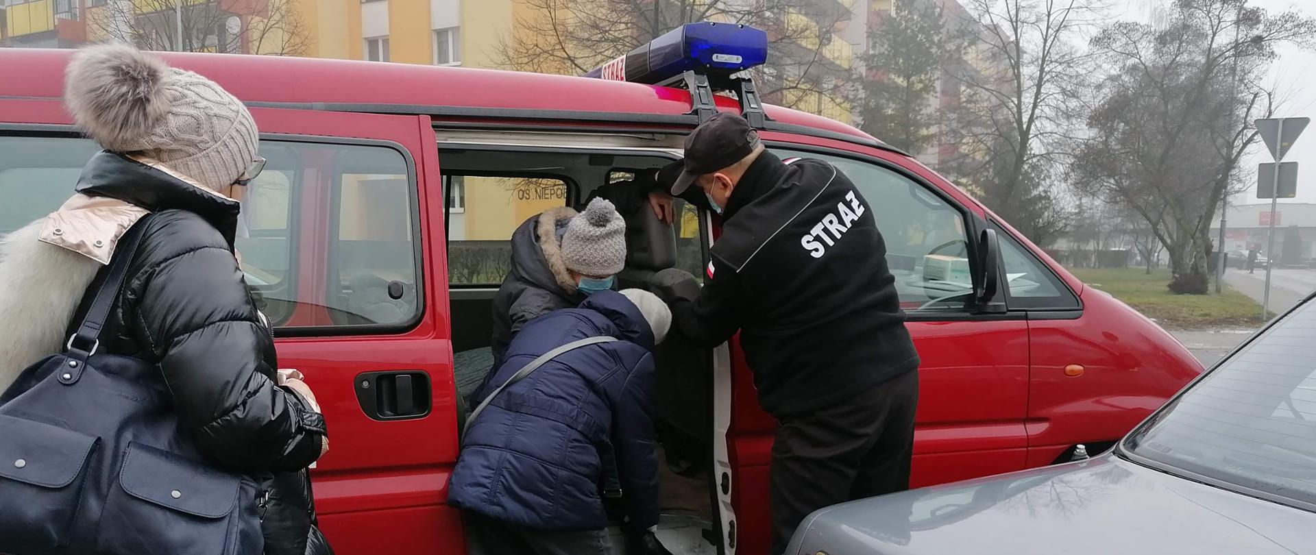 Strażak OSP pomaga wysiąść z samochodu kobiecie dowożonej na szczepienia.