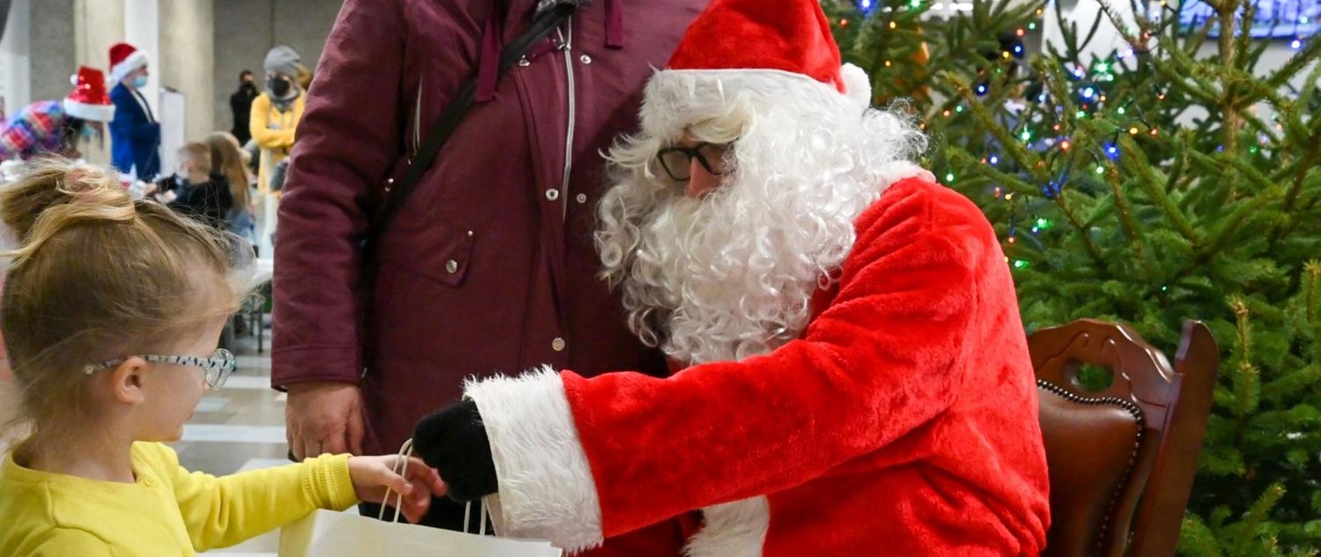 Święty Mikołaj wręcza białą torbę z narysowanymi zwierzętami z Zielonej drużyny, dziewczynce w żółtym swetrze