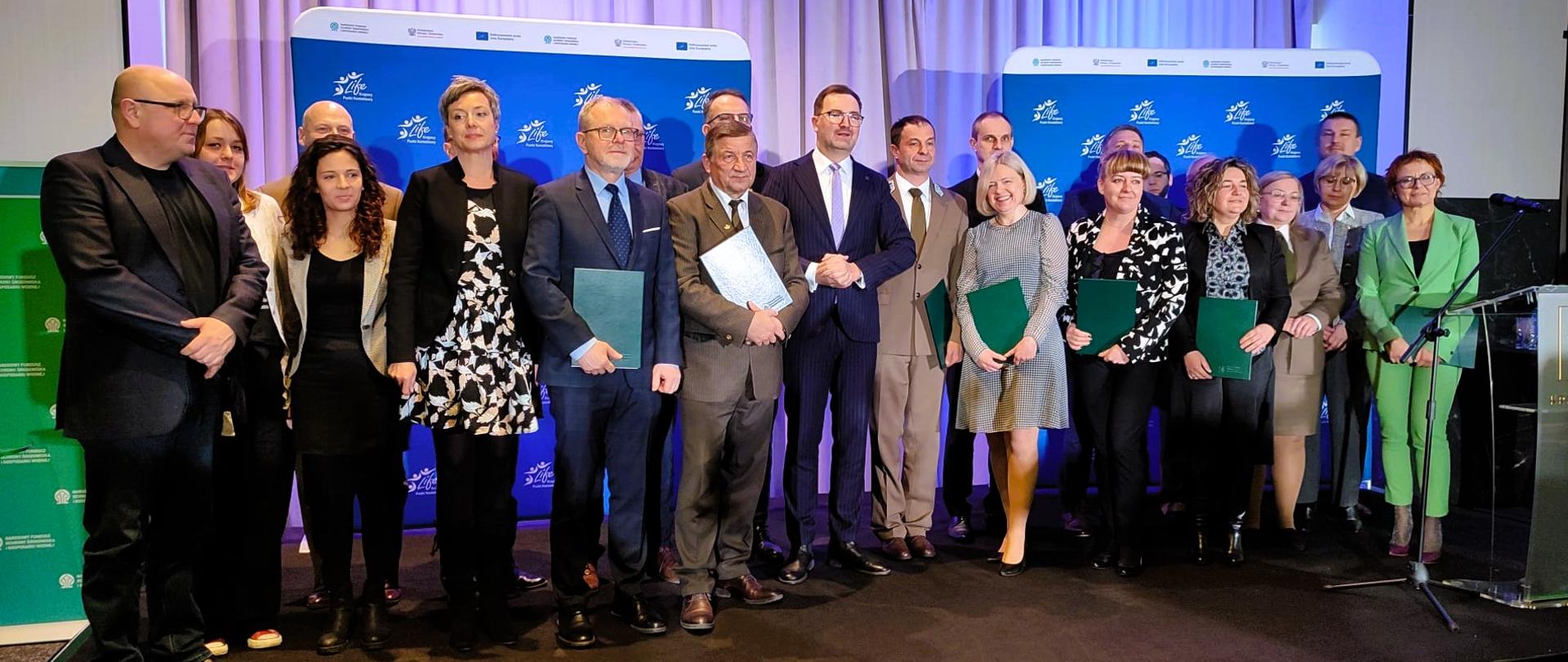 Grupa osób pozująca do wspólnej fotografii na tle ścianki promocyjnej z logiem Programu LIFE - kilka osób trzyma zielone teczki 