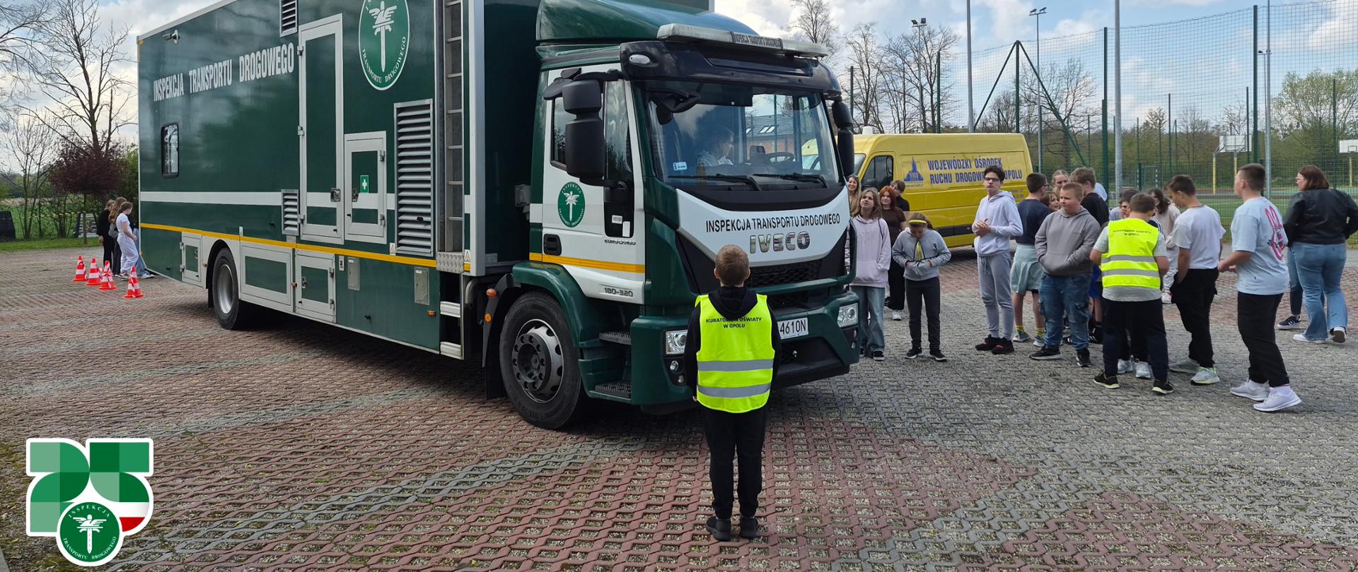 Ciężarowy pojazd Inspekcji Transportu Drogowego (IVECO) zaparkowany na placu szkolnym. Przed pojazdem stoi grupa młodzieży szkolnej oraz osoba w żółtej kamizelce odblaskowej z napisem "Kuratorium Oświaty w Opolu". W tle widoczny żółty bus Wojewódzkiego Ośrodka Ruchu Drogowego w Opolu oraz boisko sportowe.
