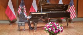 Piano recital by Jakub Kuszlik on the occasion of Poland's National Independence Day. The pianist performs works by Polish composers on a black king on stage at the Colburn School in Los Angeles. In the background are the flags of Poland and the United States. A red and white bouquet of roses stands in front of the piano. 