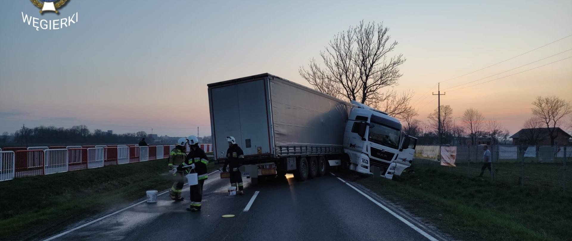 Zdjęcie przedstawia samochód ciężarowy z naczepą stojący częściowo na jezdni częściowo w rowie. Obok pojazdu stoi trzech strażaków usuwających plamę z jezdni
