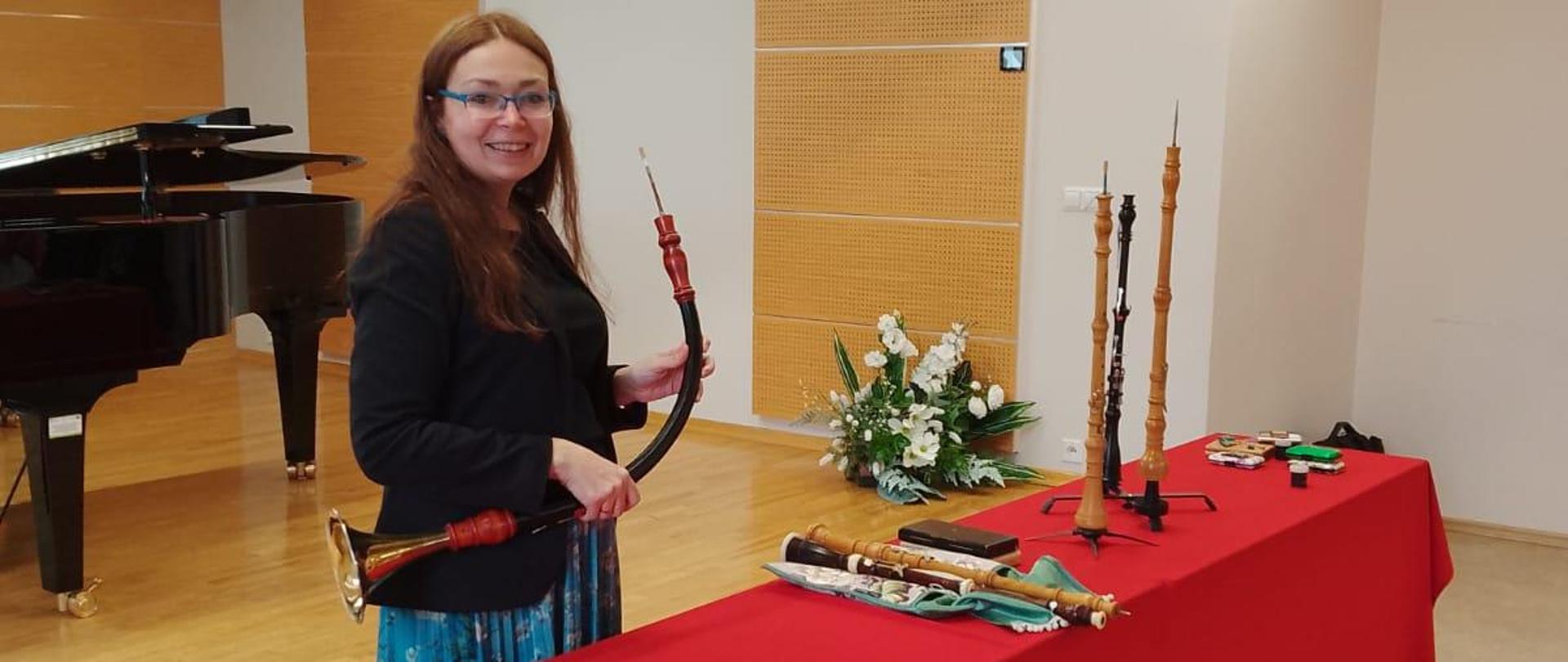 Na sali kameralnej prowadząca warsztaty przedstawia oboje barokowe. Jeden z nich trzyma w dłoni, pozostałe są położone na dwóch ławkach przykrytych czerwona płachtą. Za nią na scenie fortepian a w rogu sali sztuczna kompozycja.