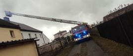 Zdjęcie przedstawia mechaniczną drabinę pożarniczą wysuniętą nad komin budynku mieszkalnego stojącą na drodze gruntowej, budynki mieszkalne i gospodarcze.