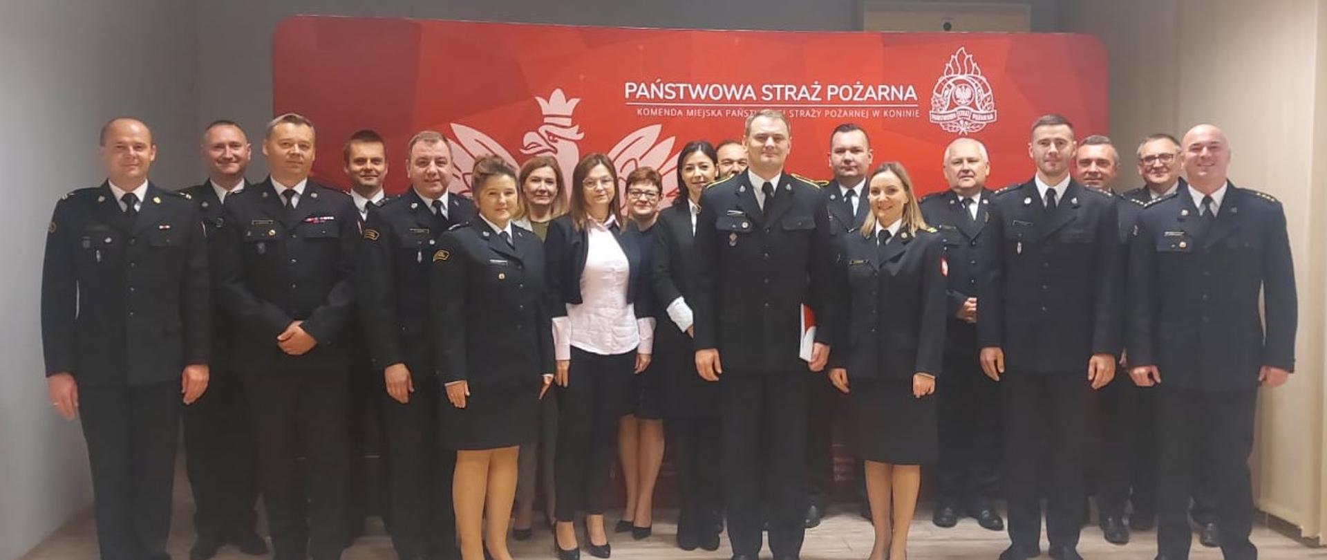 Na zdjęciu na tle ścianki Komendy Miejskiej PSP w Koninie stoi mianowany strażak, komendant, zastępca komendanta oraz funkcjonariusze i pracownicy komendy. Mianowany trzyma decyzję o mianowaniu.