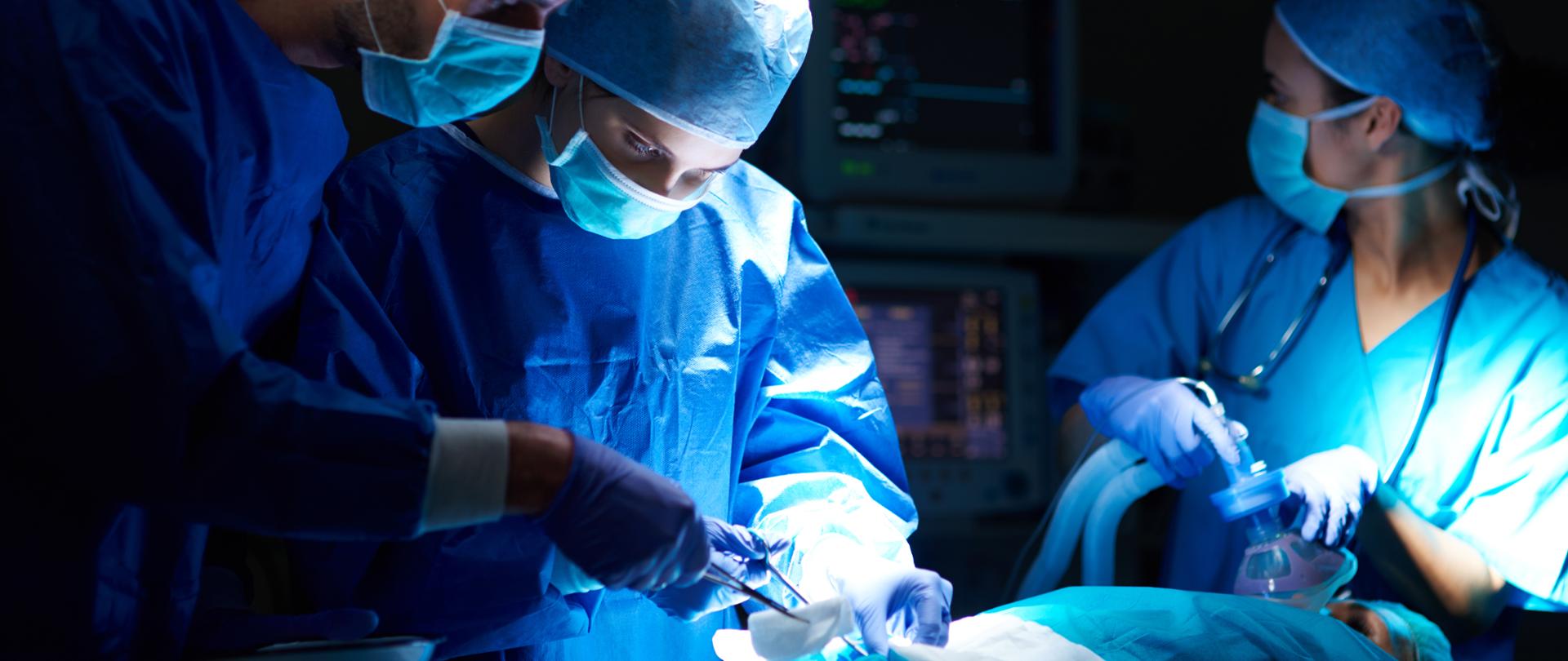 Pomieszczenie, sala operacyjna, lekarze w makach i czepkach z narzędziami w rękach pochylają się nad pacjentem leżącym na stole operacyjnym 