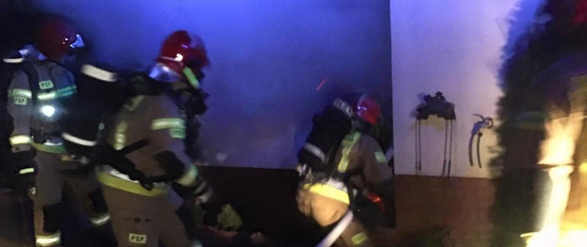 Funkcjonariusze Państwowej Straży Pożarnej próbują wejść do budynku. Każdy ze strażaków ma aparat ochrony dróg oddechowych. Pierwszy z nich ma również odcinek węża. Nad głowami strażaków unoszą się kłęby dymu. 