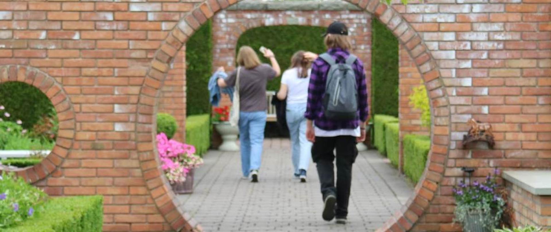 Troje ludzi odwróconych tyłem przechodzi przez okrągłą bramę z cegły