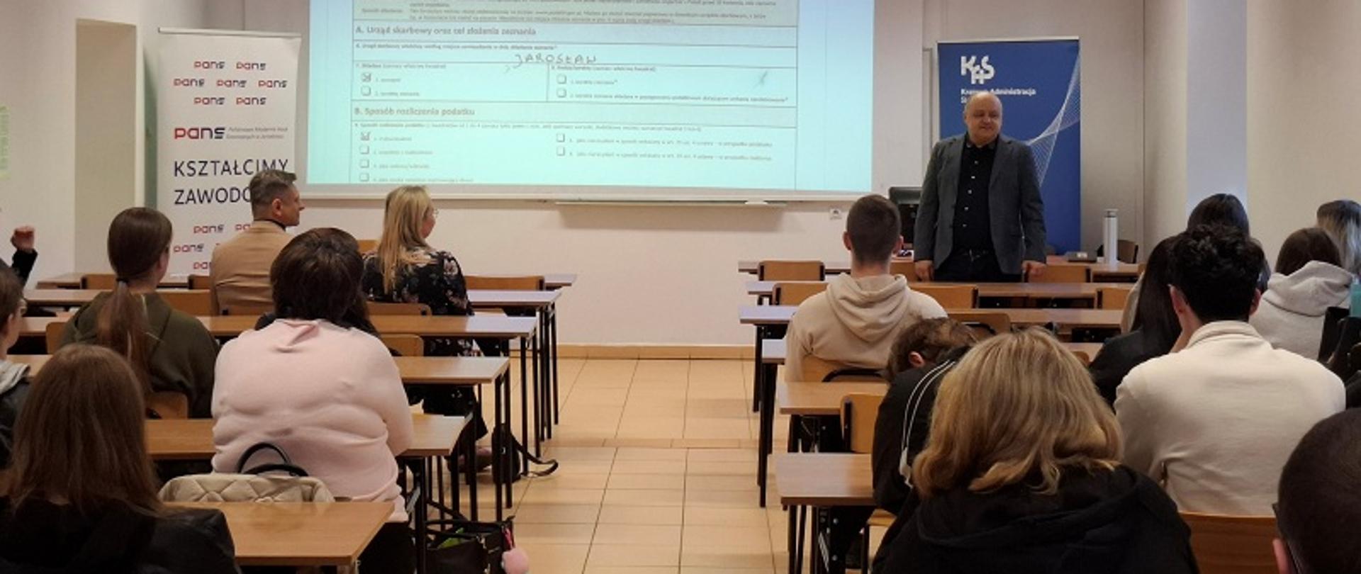 Sala wykładowa PANS. Studenci siedzą w ławkach. Przed nimi prezentację omawia pracownik urzędu skarbowego.