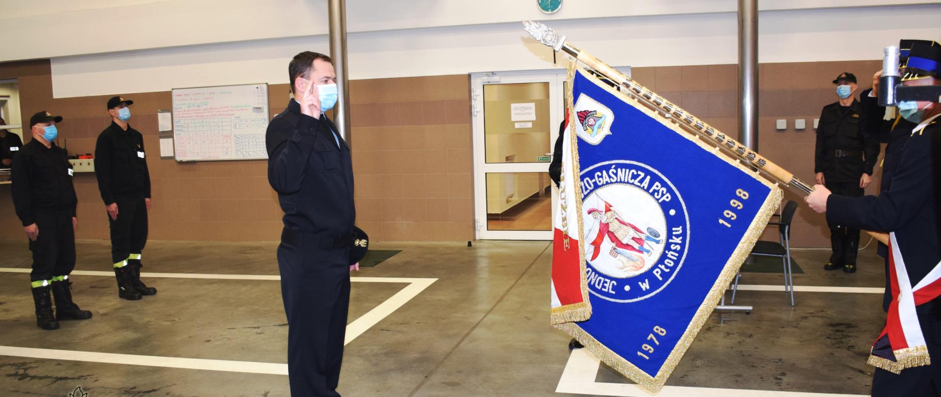 Na pierwszym planie strażak PSP, bez nakrycia głowy, z podniesioną prawą ręką do góry składa ślubowanie, z prawej poczet sztandarowy KP PSP Płońsk. Z lewej dwóch strażaków w szeregu.
