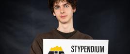 Na zdjęciu widzimy Sebastiana Zycha, ucznia kl. 5B - Stypendystę Tatrzańskiej Izby Gospodarczej 2025. Sebastian sto i w rękach trzyma tabliczkę, symboliczny czek z napisaną kwotą 7.000 zł. Na białej tabliczce dodatkowo widać logo tatrzańskiej Izby Gospodarczej i czarny napis Stypendium. Sebastian na tle czarnej ściany, jest ubrany w sweter w kolorze ciemny brąz i czarne spodnie. Na pasku w spodniach ma zawieszony klucz z czerwonym brelokiem. 
