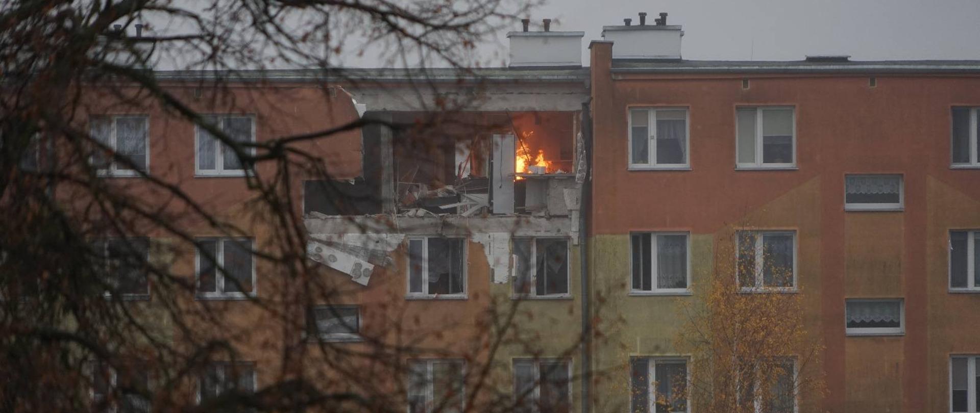 Zdjęcie zrobione z oddali. Na zdjęciu budynek mieszkalny wielorodzinny, jedno z mieszkań uszkodzone widać płomienie.