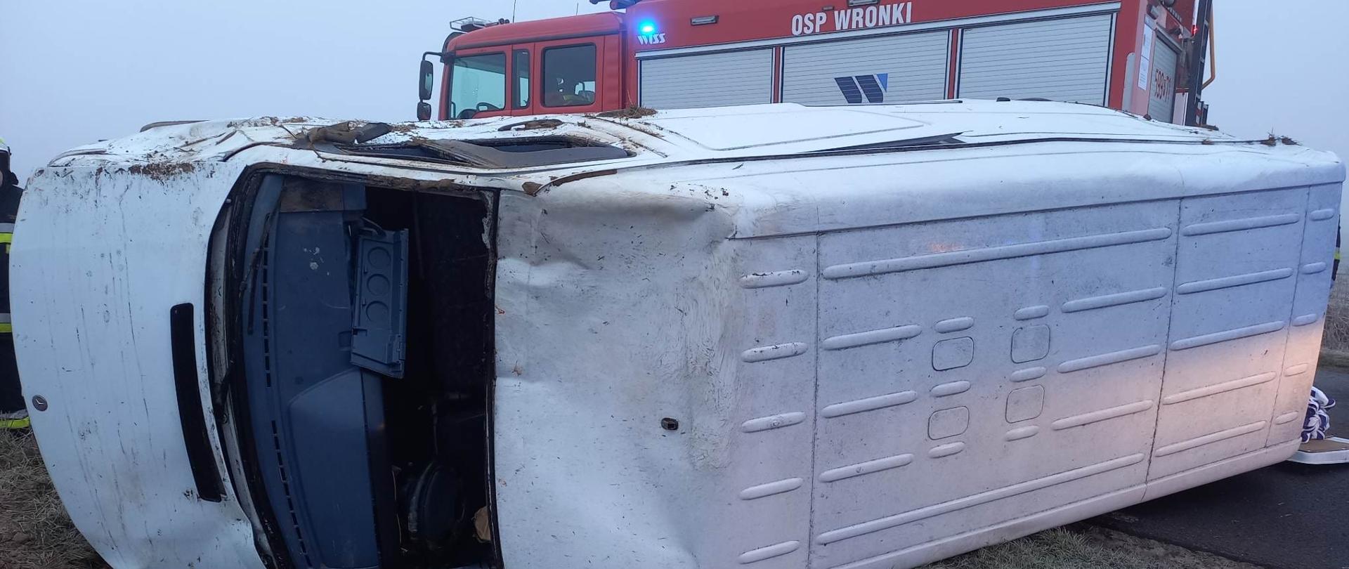 Widać leżący samochód na lewym boku, za nim samochód strażacki