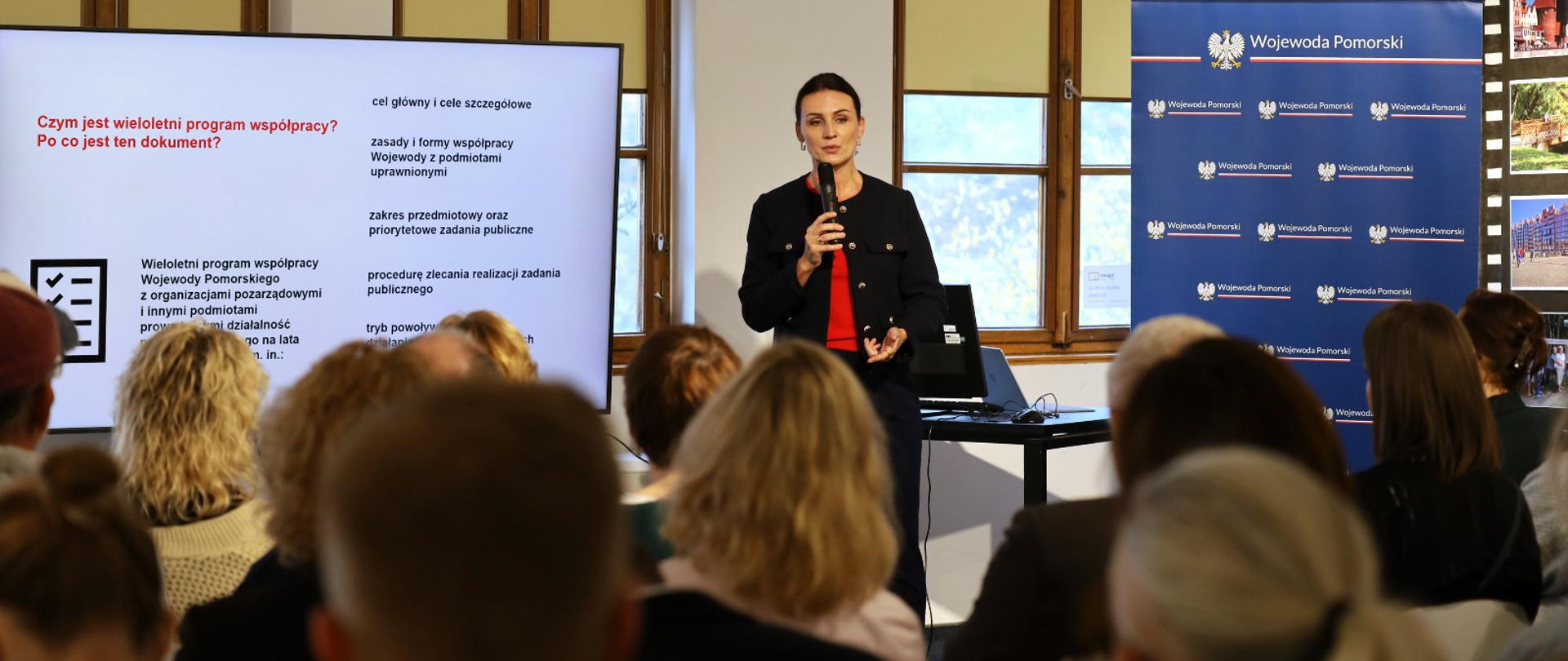 kobieta stoi z mikrofonem i przemawia do zgromadzonych, za nią tablica z napisem czym jest wieloletni program współpracy