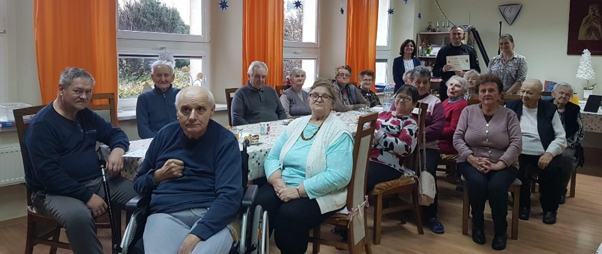 Na zdjęciu znajdują się osoby starsze siedzące przy stole, w tle widać 3 osoby