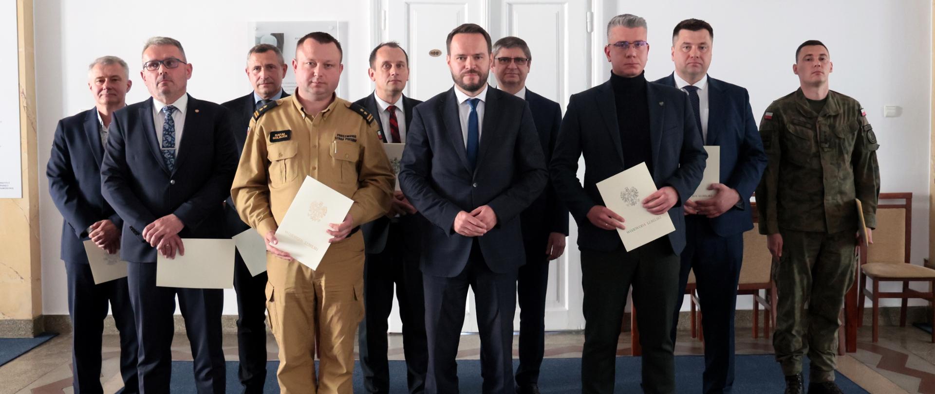 Dziewięciu mężczyzn stoi w rzędzie w sali z białymi ścianami i marmurową podłogą. W tle widoczne są drzwi z napisem "SALA IM. RYSZARDA KACZOROWSKIEGO" oraz kinkiety. Mężczyźni ubrani są w garnitury, mundury i swetry. Niektórzy trzymają w rękach białe dokumenty.
