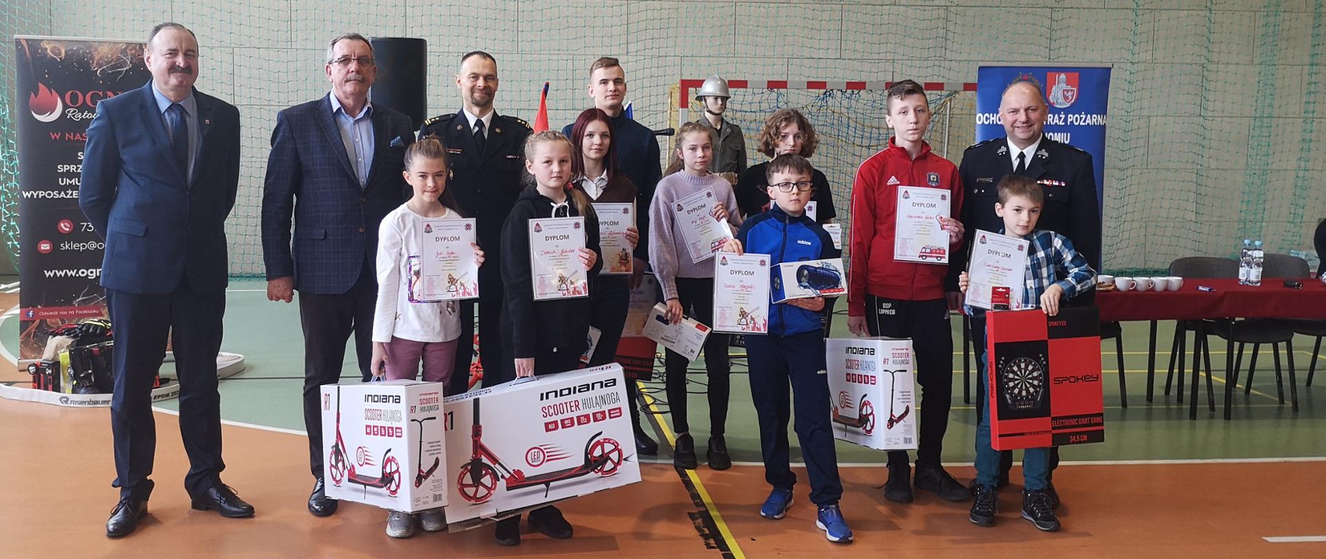 Zdjęcie zostało zrobione na sali gimnastycznej. Przedstawia zwycięzców eliminacji powiatowych OTWP. Dzieci trzymają w rękach nagrody oraz dyplomy. Za nimi stoją przedstawiciele władz oraz Komendant Powiatowy PSP w Bytowie.