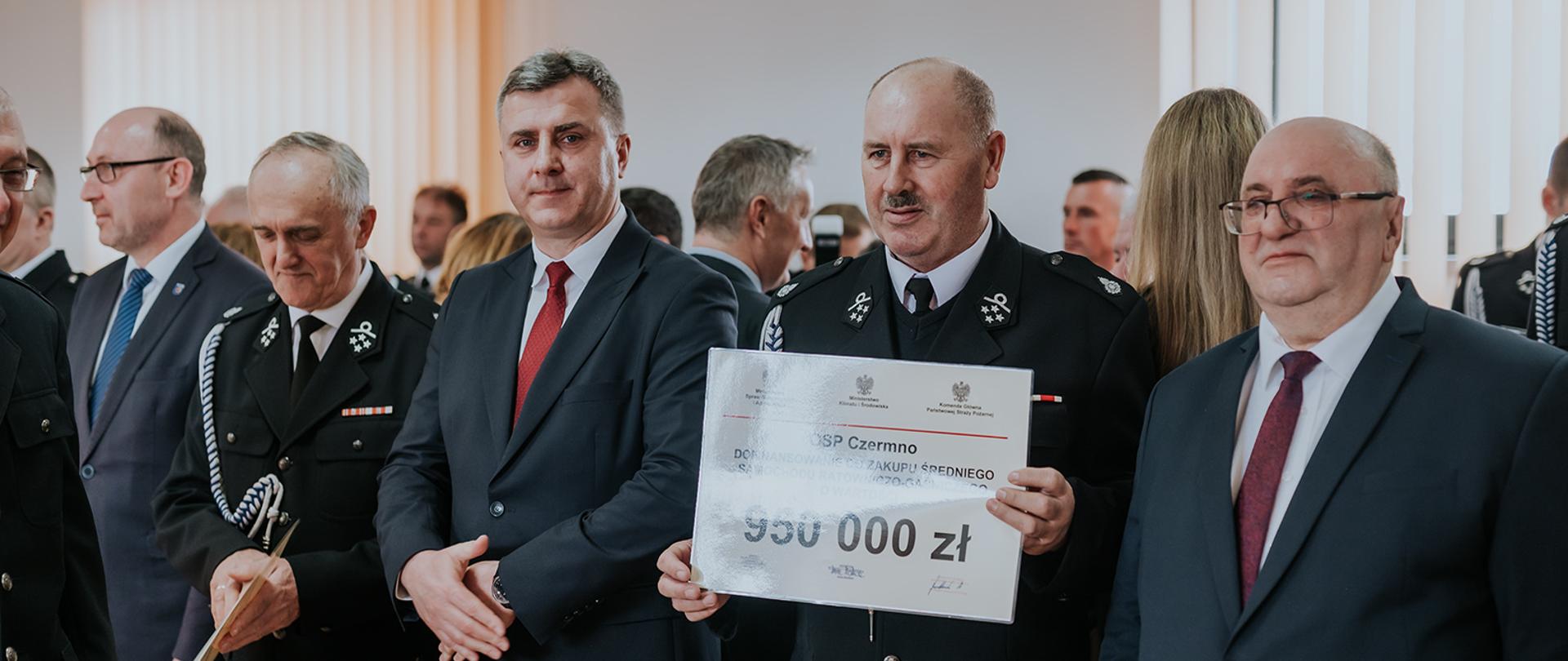 Na zdjęciu widzimy druha OSP trzymającego promesę na zakup średniego samochodu ratowniczo-gaśniczego o wartości 950 000 zł w sali podczas uroczystej zbiórki. Na blankiecie napis OSP Czermno. Na twarzach wyróżnionych uśmiechy. Strażacy OSP umundurowani w mundur galowy ze sznurem. Obok strażaka stoi mężczyzna w garniturze. 