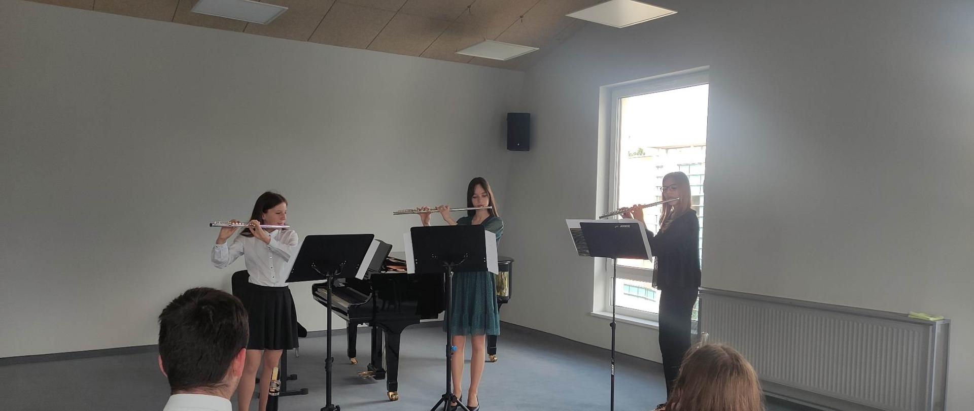 Trzy uczennice na stojąco grają na flecie, przed nimi pulpity z nutami. Za nimi okno, z grzejnik oraz fortepian i ława fortepianowa. Na pierwszym planie publiczność. Nad nimi sufit i wiszące lampy