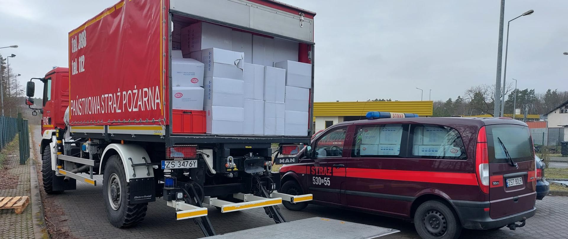 Na zdjęciu strażacki samochód ciężarowy z czerwoną plandeką załadowany kartonami, obok strażacki bus