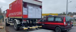 Na zdjęciu strażacki samochód ciężarowy z czerwoną plandeką załadowany kartonami, obok strażacki bus